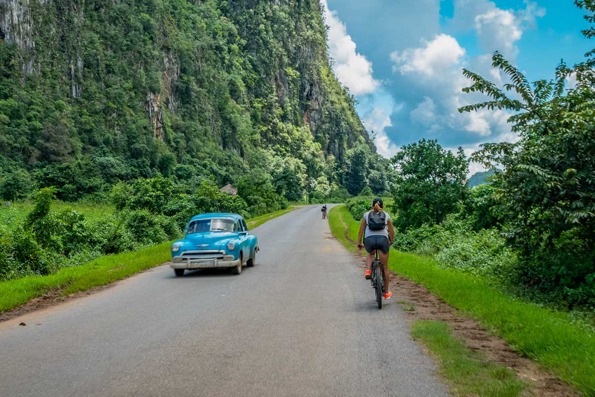 mountain biker on road with old blue car next to large mountain